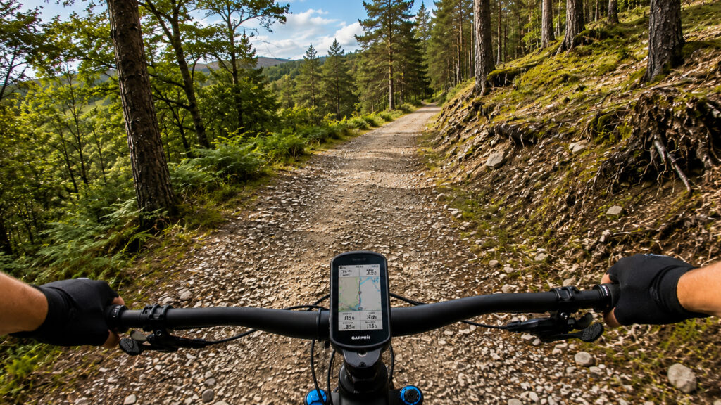 A first-person view from a mountain bike climbing a gravel fire road through woodland with a Garmin mounted on the handlebars.